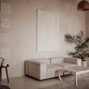Simplicity utility beauty soft hues organic shapes native eclectic textures textiles Indonesia living room to wood accents in natural light. Vivid colors palms cozy materials tropical vibes indoor potted green plants. Beige walls and niches different sizes styles waxed concrete floor large windows minimalist design ethnic. Wooden chairs at dining table in bright open space interior with sofa and abstract painting on wall.