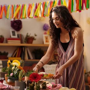 Young woman with dark long wavy hair bending over festive table and putting homemade snacks for guests while preparing for home party