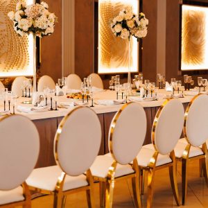 Banquet decoration in hall restaurant in golden color .Festive tables with flower composition without tablecloth and chairs for guests. Wedding set up, dinner table reception in area on party. Setting.