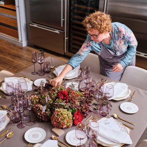 From above of curly haired woman in eyeglasses and apron standing near table with served plates and glasses while making decoration with flowers for event